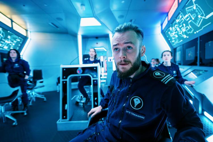 A view looking into the Bridge of the spaceship from the front. The ship is bathed in blue light. There are screens down each side, an illuminated ship's map above them, and uniformed crew members at each station.